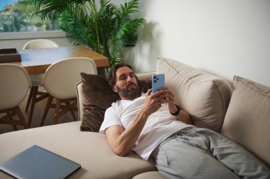 Sıradan bir adam elinde cep telefonuyla kanepeye uzanmış, teknolojiye ve rahatlığa odaklanmış temiz ve uyumlu bir evde boş vakitlerin tadını çıkarıyor..