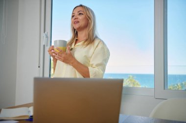 Elinde kahve fincanı tutan memnun bir kadın, elinde dizüstü bilgisayarla bir masanın başında duruyor, ofisinin penceresinden okyanus manzarasının keyfini çıkarıyor..
