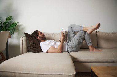 Bej bir kanepeye uzanmış şık ve rahat bir oturma odasında akıllı telefon kullanan sıradan giysili bir adam. Sahne rahatlama, modern teknoloji kullanımı ve minimalist bir yaşam tarzı içeriyor..