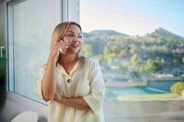 Neşeli bir kadın büyük bir pencerenin yanında duruyor, canlı bir telefon görüşmesinden zevk alıyor. Geniş oda ve manzaralı yeşillik, sakin ama profesyonel bir atmosfer ekler..