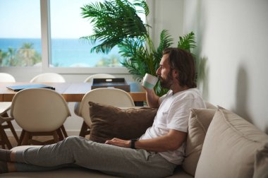 Bir adam minimalist dekorlu, parlak, rahat bir odada kanepede kahvesini içmeyi sever. Camdaki sakin kıyı manzarası rahatlatıcı atmosferi arttırıyor, dinlenmek için mükemmel..