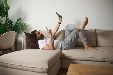 Bir adam bej bir kanepede uzanır, akıllı telefonunu gündelik ortamında kullanır. Oda modern bir dekor ve sakin bir ortam içeriyor, rahatlama, rahatlama ve bağlantıyı vurguluyor..