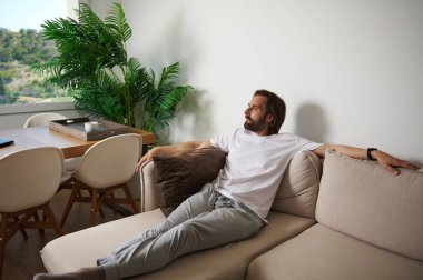 Evdeki bir adam, bej renkli bir koltukta, doğal ışıkla dolu minimalist bir oturma odasında, rahatlatıcı bir atmosfer sergiliyor..