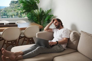 Bir adam bej bir kanepede dizüstü bilgisayarıyla oturuyor, uzaktan kumandayla meşgul. İç mekanda modern mobilya ve yeşillik, rahatlatıcı bir çalışma alanı yaratıyor..