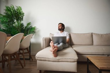 Modern mobilyaların, canlı yeşil bir bitkinin ve doğal ışığın bulunduğu şık bir oturma odasındaki koltukta uzaktan çalışan bir adam..