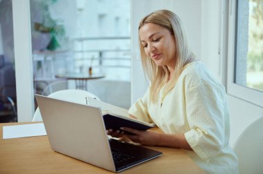 Minimalist bir ofiste masasında uzaktan çalışan bir kadın dizüstü bilgisayar kullanırken defter okuyor. Sahne üretkenlik, odak ve rahat bir çalışma ortamı sunuyor..