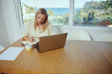 Bir kadın dizüstü bilgisayar ve defterle bir masada oturuyor, minimalist, sakin manzaralı bir yerde evden çalışıyor. Uzaktan çalışma, üretkenlik ve yaşam tarzı dengesini temsil eder.