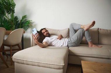 Modern bir oturma odasındaki bej bir kanepede elinde akıllı bir telefonla rahat bir şekilde uzanan bir adam. Dekor parlak, minimalist ve kapalı yeşil bitkiler içeriyor..