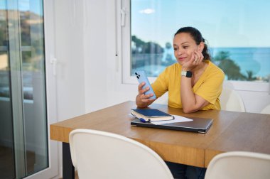 Ofisindeki bir masada oturan bir kadın akıllı telefonuna düşünceli bir şekilde bakıyor. Oda sakin bir plaj zeminine sahip. Uzaktan çalışma ve eğlence arasındaki dengeyi vurguluyor..