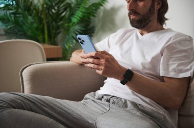 Kanepede oturan adam akıllı telefon kullanıyor, modern bir evde günlük kıyafetler giyiyor. Canlı bitkiler rahat ve uzak çalışma için sakin ve modaya uygun bir atmosfer yaratırlar..