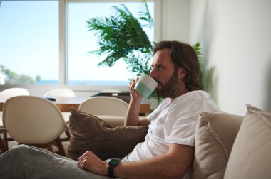 Bir adam modern, minimalist bir evde koltuğa rahatça oturur ve bir fincan kahvenin tadını çıkarır. Parlak ve havadar alan huzurlu ve sakin bir atmosfer yaratır..