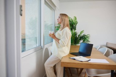 Bir kadın elinde kahve fincanıyla pencerenin kenarında duruyor ve ahşap masada dizüstü bilgisayarı ile bir dakikalık sakinliğinin tadını çıkarıyor..