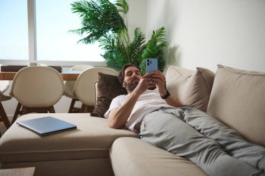 Kanepeye yaslanmış bir adam akıllı telefonunu doğal ışık alan parlak, minimalist bir oturma odasında kullanır. Dijital temas için mükemmel, rahat ve modern bir atmosfer yaratır..