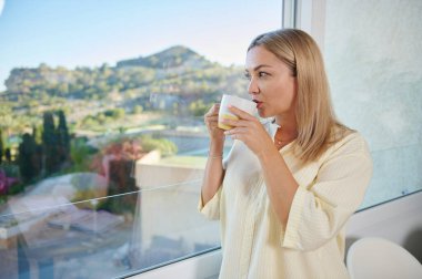 Bir kadın evde rahatlar, içkisini yudumlar ve pencereden manzara manzarasını seyreder. Bu an, doğanın güzelliği için sükunet ve takdir uyandırır..