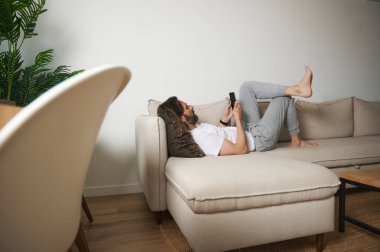 Bir adam rahat bir şekilde rahat bir şekilde kanepede uzanır ve akıllı telefonuyla meşgul olur. Huzurlu yaşam alanı, rahat atmosfere eklenen minimalist dekora sahiptir..
