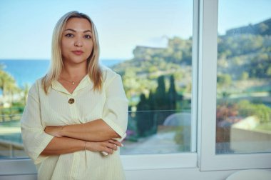 Bir kadın kendine güvenerek geniş bir pencerenin yanında duruyor, bereketli manzaranın ve uzak denizin huzurlu manzarasının tadını çıkarıyor, zarafet, sükunet ve modernliği birleştiriyor..