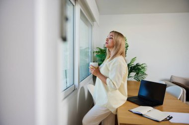Doğal elementler ve dizüstü bilgisayarla süslenmiş, dengeli çalışmayı ve gevşemeyi sembolize eden minimalist bir ev ofisinin penceresinin yanında kahvesini yudumlayan bir kadının huzurlu anı..