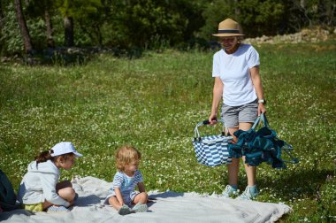 Doğada sakin bir aile, anneleri pikniği hazırlarken battaniyeye oturmuş çocukları tasvir ediyor, çevresi yeşillik ve güneşle çevrili, dış mekandaki ahengi somutlaştırıyor..