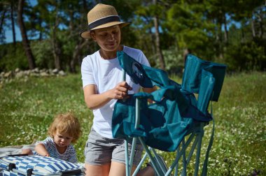 Bir anne, çocuğu canlı ağaçlarla çevrili güneşli bir çayırda oynarken sandalyeye yardım eder..