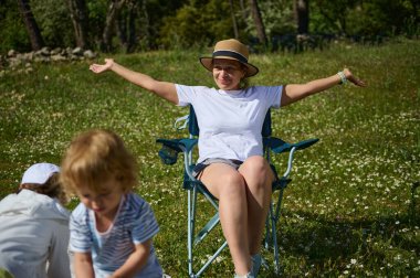 Neşeli bir kadın papatya tarlasında dinleniyor, çocuklar dışarıda eğlenirken, yeşilliklerle çevrili güneşli bir piknik atmosferinin tadını çıkarıyor..