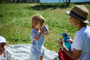 Güneşli bir günde yemyeşil bir parkta piknik yaparken anne ve çocukların iyi vakit geçirdiği iç açıcı bir açık hava sahnesi..