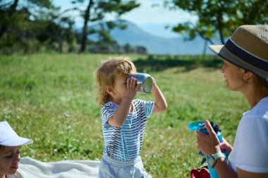 Güneşli bir günde güzel bir dağ manzarasının ortasında aile pikniği. Bir çocuk bardaktan içerken anne sevgi dolu bir şekilde dış mekanın neşesini somutlaştırıyor..