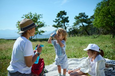 Aile, dışarıda kaliteli zaman geçirir, pikniğin tadını çıkarır. Etrafı yeşillik ve güneş ışığıyla çevrilidir. Birlikteliği ve doğal çevreye olan sevgiyi gösterir, bağlantıyı ve açık havadaki keyfi vurgular..