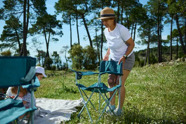 Bir anne ve çocukları güzel bir orman çayırında güneşli bir pikniğin tadını çıkarıyorlar. Küçük sandalyeler ve battaniye kuruyorlar. Aile bağı kurmak ve doğanın tadını çıkarmak için mükemmel anlar..