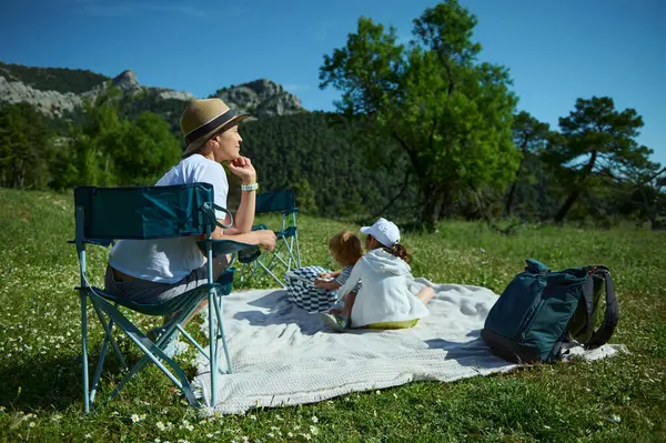 Mutlu bir anne ve çocukları piknik sırasında dağlarla, yemyeşil ağaçlarla ve parlak mavi gökyüzüyle çevrili bir çayırda birbirlerine bağlanıyorlar..