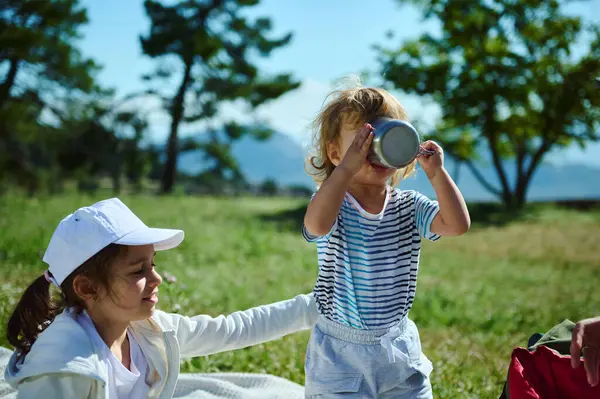 İki küçük çocuk parkta güneşli bir piknik gününün tadını çıkarıyor. Bir çocuk metal bardaktan yudumlarken, diğeri sıcak bir şekilde gülümsüyor, neşe ve aile bağlarını çağrıştırıyor..