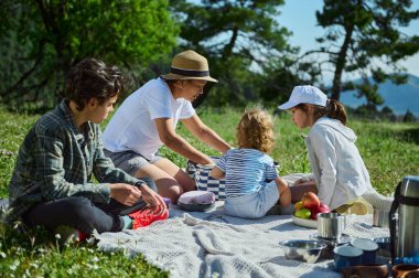 Bir anne ve çocukları, manzaralı dağ manzaraları ve yemyeşil yeşilliklerle çevrili rahatlatıcı bir pikniğe katılıyorlar. Ulusal Piknik Ayı 'nın neşesini somutlaştırıyorlar..