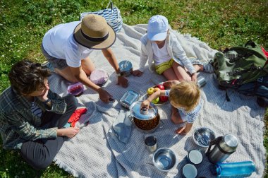 Doğada güneşli bir pikniğin tadını çıkaran bir aile. Birlikte bir battaniyeye oturup, yemeklerini paylaşıyorlar ve ılık yaz günlerini kutluyorlar..