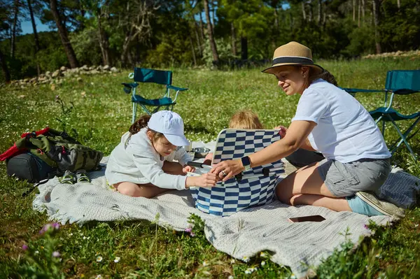 Bir anne ve çocukları huzurlu bir dağ çayırında keyifli bir pikniği paylaşıyor, güneşli havanın tadını çıkarıyor ve aile bağlarını güçlendiriyor..