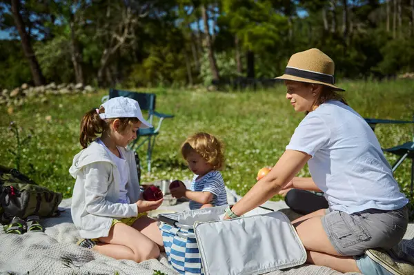 Bir anne ve çocukları yemyeşil doğada keyifli bir pikniği paylaşıyor, güneşli havanın ve aile bağlarının tadını çıkarıyorlar. Canlı sahne neşe, sükunet ve unutulmaz anları vurguluyor..