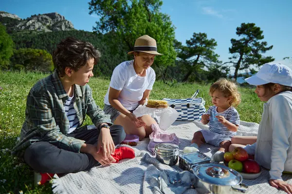 Anne ve çocukları olarak sıcacık bir aile piknik yapmak için güzel dağ manzarası ve yaz güneşinin altında canlı yeşillikler arasında toplanıyor..