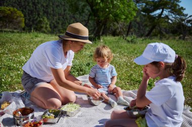 Bir anne ve çocukları güneşli bir günde manzaralı bir dağ manzarasında keyifli bir açık hava pikniğini paylaşıyorlar..