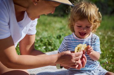 Bir anne ve küçük çocuğu güneşli bir yaz gününde keyifli bir açık hava piknik deneyimini paylaşıyorlar, doğa ile çevrili, bağ kuruyor ve lezzetli bir yemeğin tadını çıkarıyorlar..