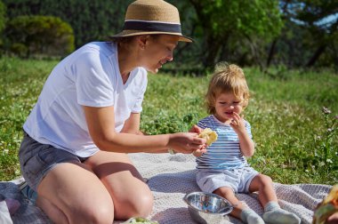 Bir kadın ve küçük çocuğu açık havada keyifli bir piknik sahnesi paylaşıyorlar. Sıcak ve güneşli bir gün dingin dağ zeminini güçlendirir. Aile bağlarını ve doğaya olan sevgiyi vurgular..