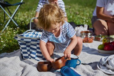Küçük bir çocuk, güneşli bir aile ortamında çim çayırında toplanan piknik eşyalarıyla meşgul olur. Bu, doğanın neşesini, bağlantısını ve değerini sembolize eder..