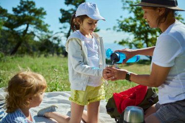 Bir anne, dağlarda güneşli bir piknik sırasında çocuklarına içki koyar. Aile bağlarını, açık hava eğlencelerini ve doğanın güzelliğini vurgular..