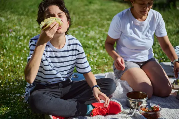 Bir aile güzel güneşli bir günü yemyeşil bir açık hava ortamında pikniğin tadını çıkararak geçirir. Çocuk neşeli bir ifadeyle taco yerken anne piknik düzenlemelerine yardım ediyor..