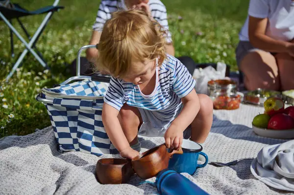 Küçük bir çocuk, güneşli bir aile ortamında çim çayırında toplanan piknik eşyalarıyla meşgul olur. Bu, doğanın neşesini, bağlantısını ve değerini sembolize eder..