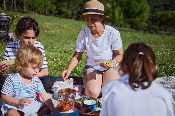 Bir anne ve çocuklar pikniği güzel bir dağ manzarasında paylaşıyorlar. Açık hava yemekleri sofrası, güneşli bir yaz gününde aileyi, birlikteliği ve doğanın tadını çıkarmayı ön plana çıkarır..