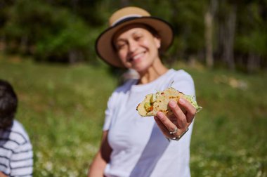 Dışarıda mutlu bir şekilde vakit geçiren bir kadın piknik sırasında taco uzatıyor. Bereketli yeşil ortam ve güneşli atmosfer boş zamanları, birlikteliği ve açık hava yemeklerini vurguluyor..