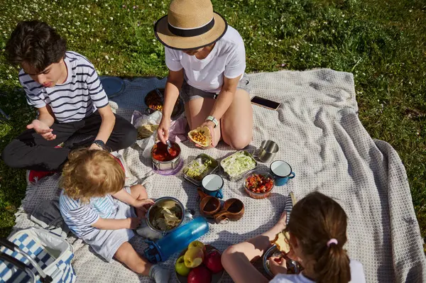 Dört kişilik neşeli bir aile piknik battaniyesinde oturup tako yiyerek kaliteli zaman geçiriyorlar. Manzaralı çevre ve güneşli hava birlikteliği ve aile bağlarının neşesini vurguluyor.