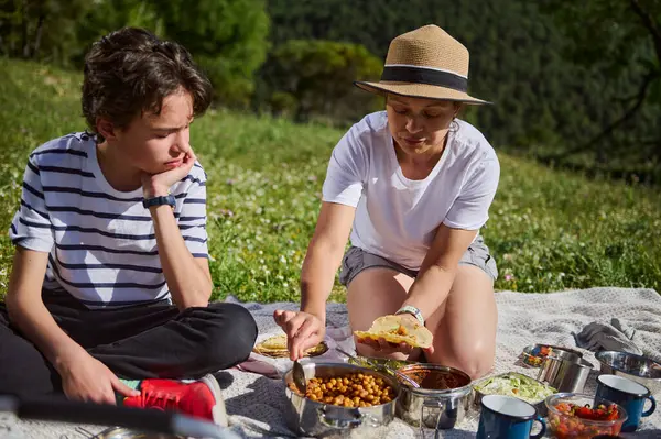 Neşeli bir aile doğada pikniğin tadını çıkarır, dağlarla çevrili bir battaniyenin üzerinde oturur, lezzetli tacoları paylaşır ve tadını çıkarır. Sahne beraberliği ve açık havanın takdir edilmesini gösteriyor..