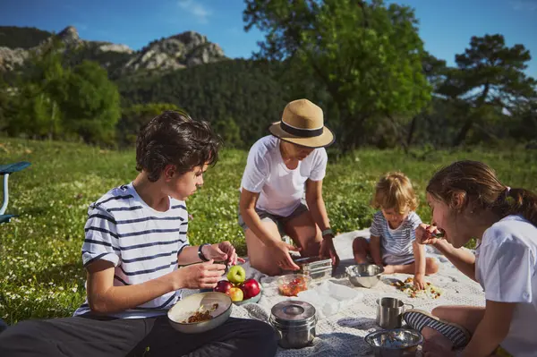 Bir anne ve çocukları güzel bir açık hava pikniğini paylaşıyorlar. Resimli bir dağ manzarasının ortasında, aile bağlarını, paylaşılan anları vurgulayarak ve doğanın güzelliğini kucaklayarak..