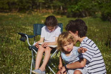Anne, çocuklarıyla yemyeşil ve manzaralı bir ortamda rahatlatıcı bir günün tadını çıkarıyor. Birlikte keyifli anlar paylaşıyorlar, açık havada sevgi ve beraberlik sergiliyorlar..
