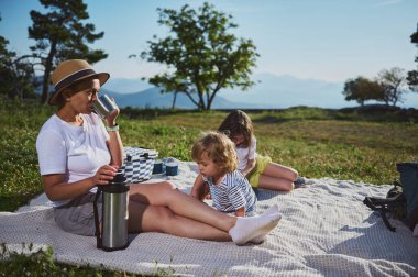 Anne ve çocuklar dağlarda piknik yaparken dışarıda kaynaşıyorlar. Görüntü, güzel bir doğal ortamda aile birliği, rahatlama ve doğanın tadını çıkarıyor..