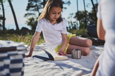 Genç bir kız, güneşli bir açık hava toplantısında piknik battaniyesinde dinlenirken kitap okumaktan zevk alır. Doğada neşe ve rahatlığın vücut bulmuş halidir..